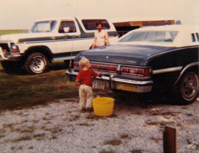 White Ford Bronco behind a green Ford sedan being washed by a man and toddler with yellow bucket, Sept 1981.