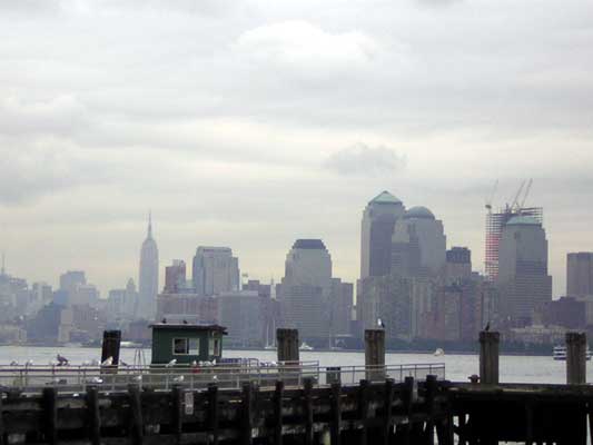 New York City skyline with Empire State Building and World Trade Center towers under a cloudy sky, viewed from a pier