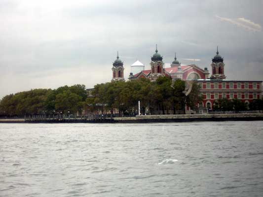 Building with red and white facade and domed towers behind trees, viewed across choppy water under a cloudy sky