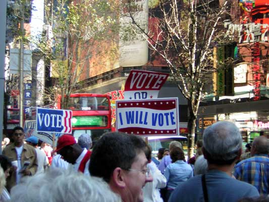 People gathered on a city street with signs that say "VOTE" and "I WILL VOTE". A red double-decker bus is visible
