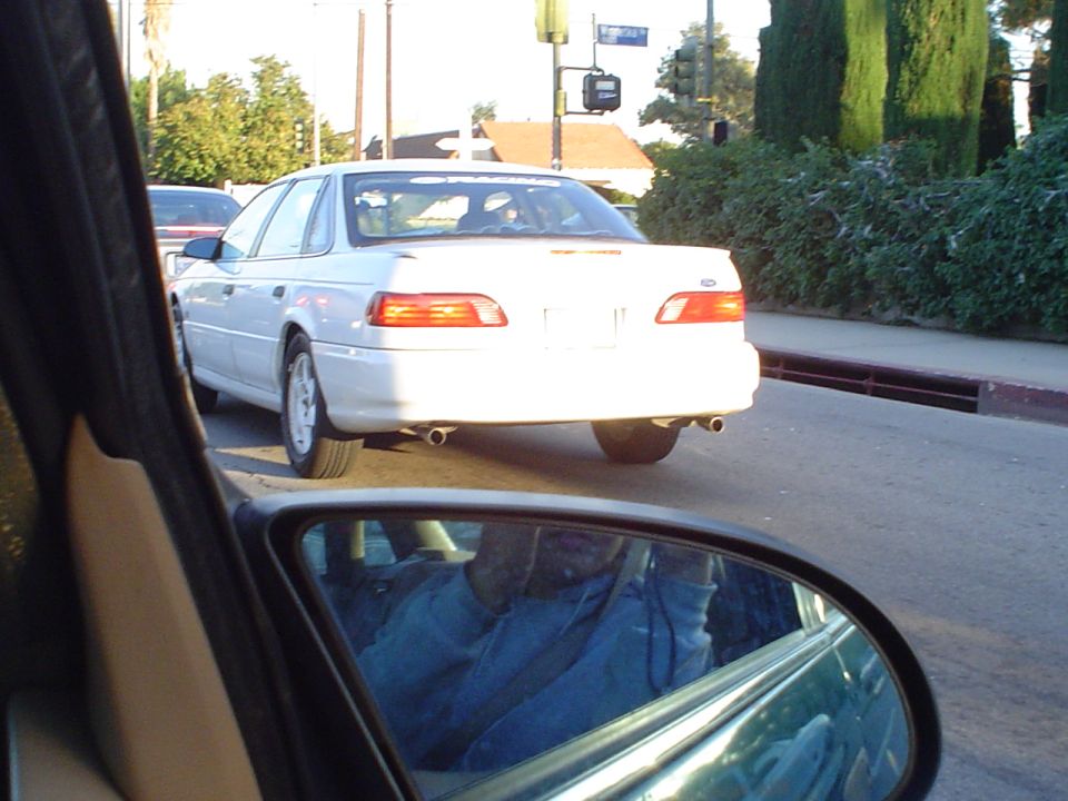 White sedan from the rear, seen from a car's side mirror. The car has dual exhaust pipes and a visible Ford emblem