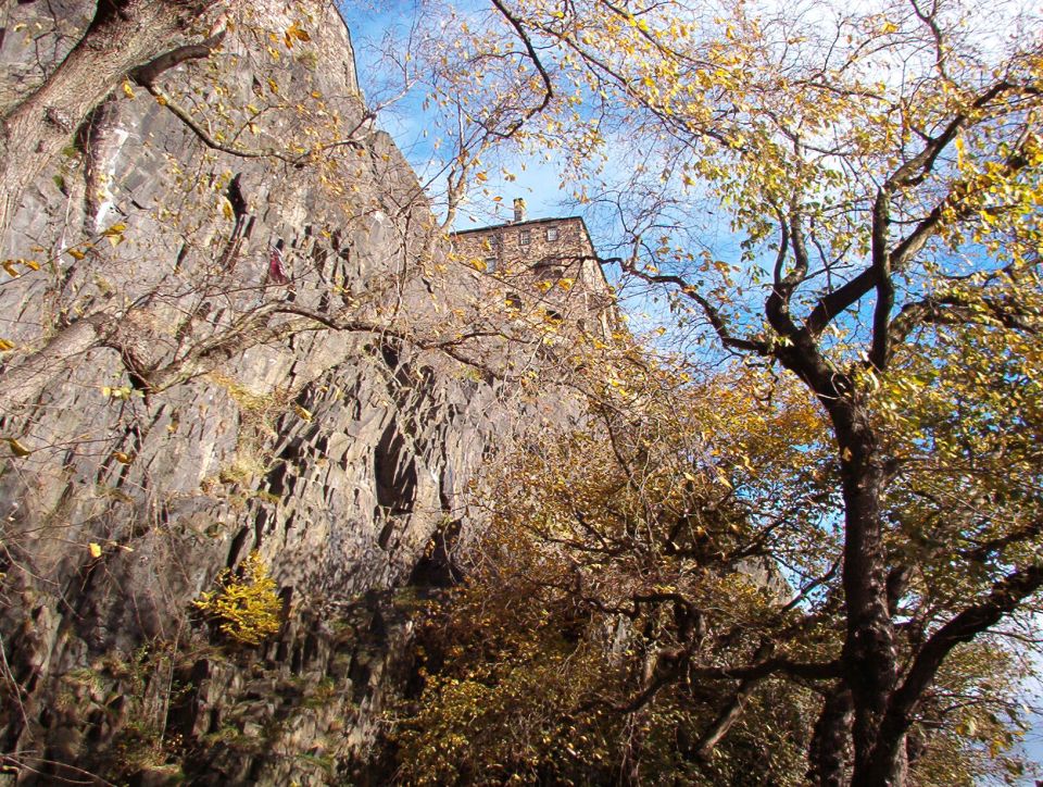 Rocky cliff and autumn trees with a stone building against blue sky.