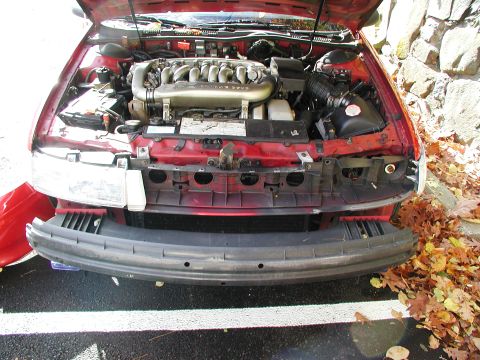 1992 Ford Taurus - The engine bay of a red car is visible, with the front bumper detached and lying on the ground.