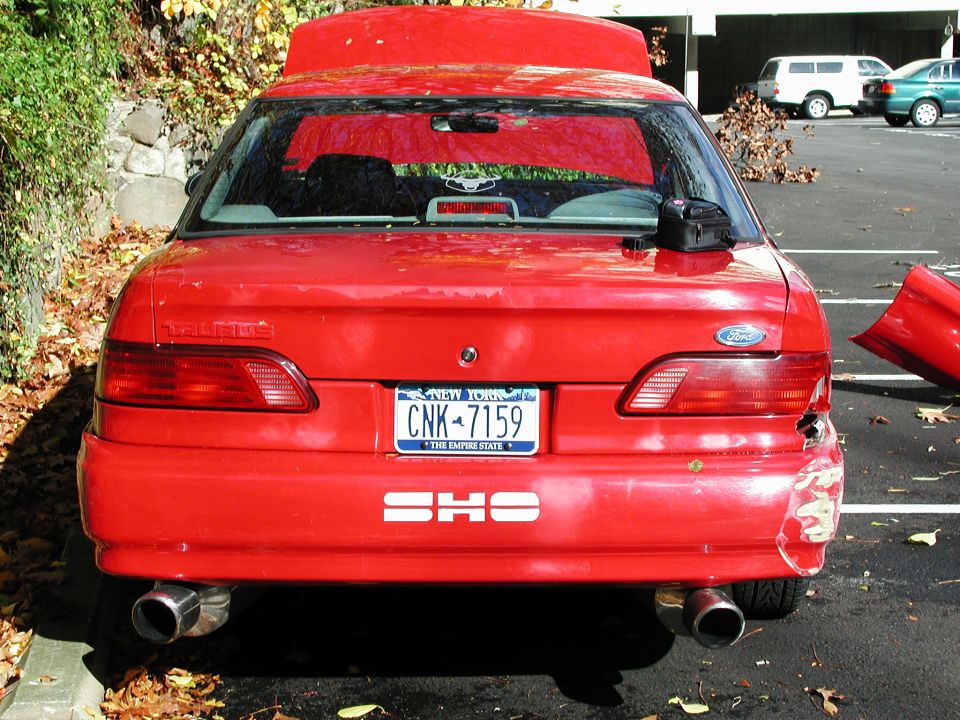 Rear view of a red 1992 Ford Taurus SHO with damage to the right rear bumper and taillight. A detached bumper piece is visible to the right