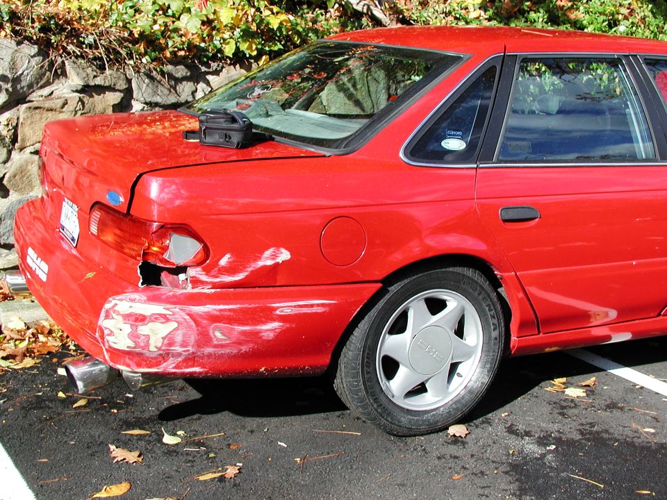red sedan, looks like a 1992 Ford Taurus, with damage to the rear bumper and taillight