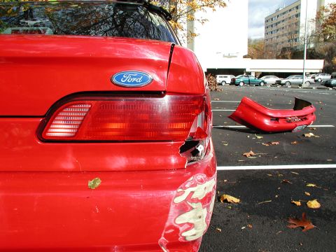 1992 Ford Taurus - The rear bumper of a red Ford sedan lies on the ground next to the vehicle after sustaining damage.