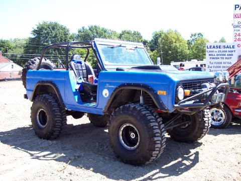 1974 Ford Bronco - A bright blue early Ford Bronco with the top removed, shown from a 3/4 front angle on a gravel lot. It has a black roll cage, oversized mud…