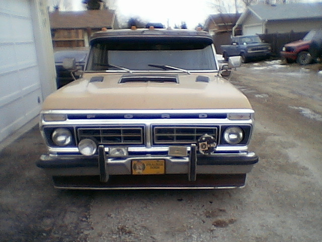 Front view of a tan Ford pickup with FORD grille lettering, hood scoop, and bumper-mounted driving lights on gravel driveway