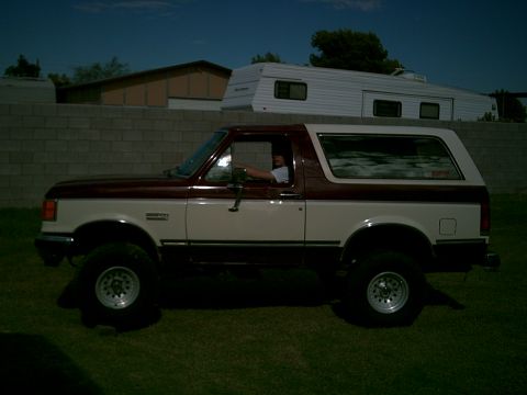 1988 Ford Bronco - A two-tone maroon and white Ford Bronco is parked on a grassy area in front of a brick wall and a trailer.