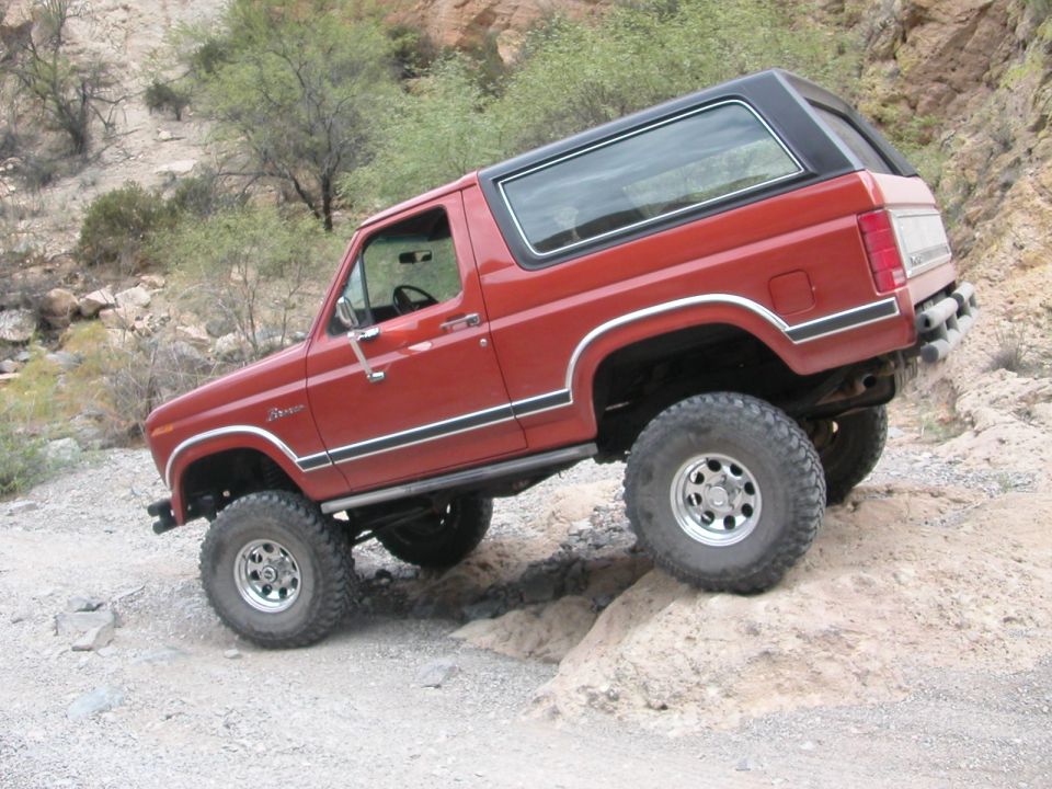 Red 1984 Ford Bronco with lifted suspension and off-road tires navigating a rocky incline