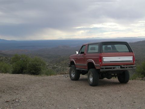 1984 Ford Bronco - This lifted Ford Bronco is parked on a dirt road overlooking a vast, hilly landscape under a cloudy sky.