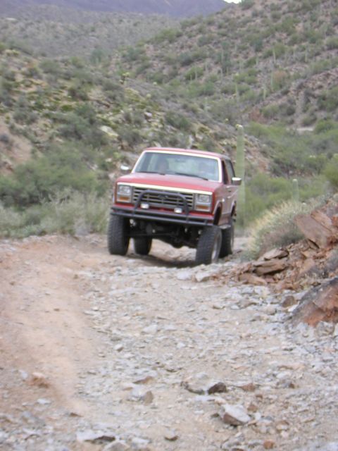 1984 Ford Bronco - A red SUV navigates a rough, rocky path through a desert environment with sparse vegetation.