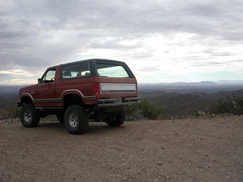 1984 Ford Bronco - Red Ford Bronco with lifted suspension parked on a dirt road overlooking a mountainous landscape.