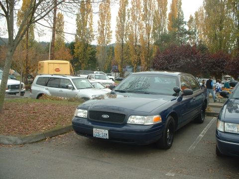1999 Ford Crown Victoria - A dark blue sedan is parked in a lot with other vehicles and trees in the background.