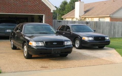 1999 Ford Crown Victoria - Two black Ford Crown Victorias are parked in a driveway in front of a brick garage and house.