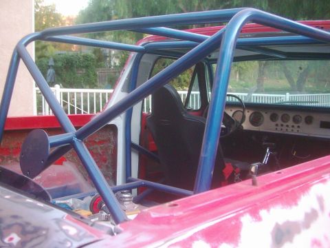 1978 Ford Bronco - Blue-painted full roll cage installed in a topless red early Bronco, viewed from the rear bed looking forward into the cab. A black high-ba…