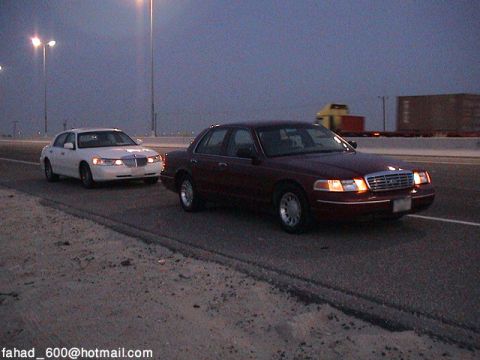 2000 Ford Crown Victoria - Two Ford Crown Victorias, one maroon and one white, travel down a highway as twilight descends.