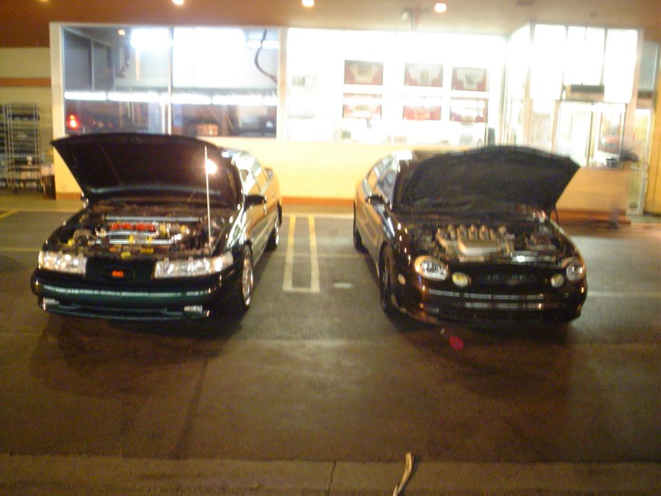 Two dark sedans parked side-by-side, both with hoods open showing their engines
