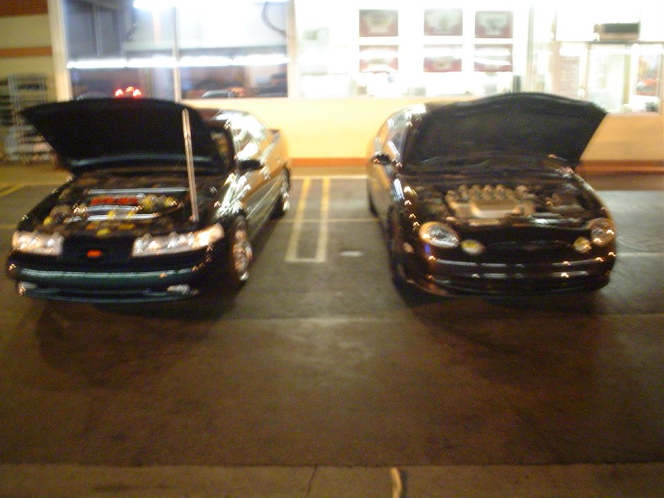 Two dark sedans parked side-by-side, hoods open, showing engines