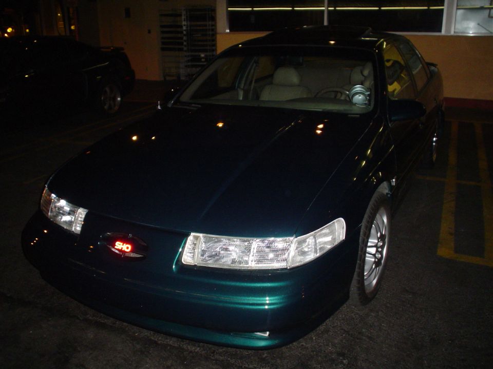 Dark green sedan with a red SHO badge on the grille. Another dark vehicle is parked to the left