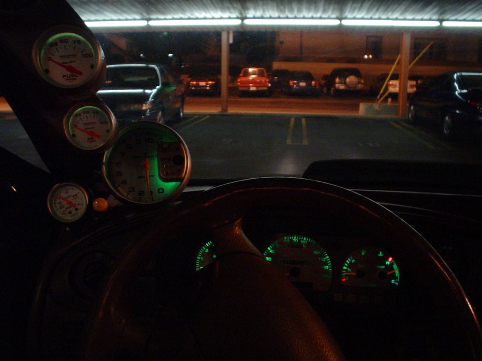 Car interior at night, showing a steering wheel, dashboard gauges, and aftermarket gauges on the A-pillar. Other cars are visible in the parking lot outside