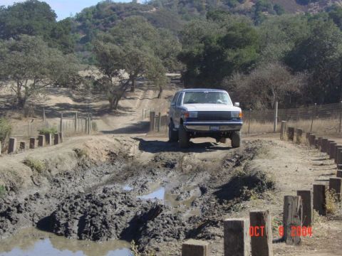 1990 Ford Bronco - A white 1990 Ford Bronco tackles a challenging muddy path on a sunny day.