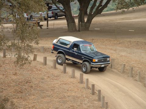 1990 Ford Bronco - 1990 Ford Bronco navigating a dirt road with trees and other vehicles in the background.