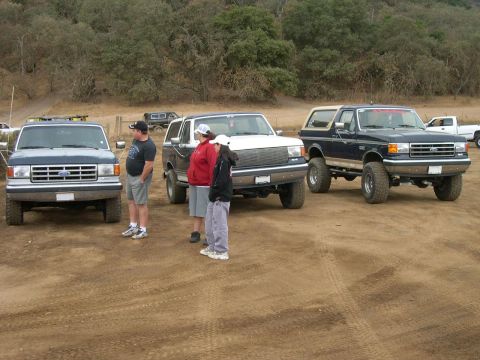 1990 Ford Bronco - Several Ford Bronco SUVs are parked on a dirt lot with trees in the background.