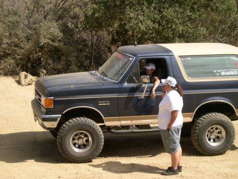 1990 Ford Bronco - A Ford Bronco with off-road tires is parked on a dirt road with two people near it.