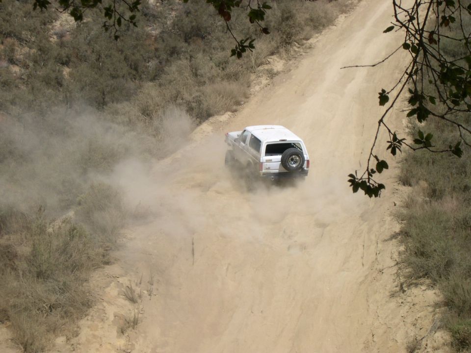 White 1990 Ford Bronco driving on a dusty dirt road, rear view