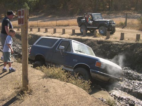 1990 Ford Bronco - A blue Ford Bronco is stuck in a mud hole while a Jeep watches from higher ground.
