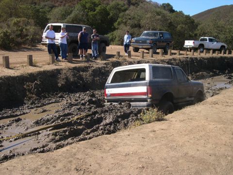 1990 Ford Bronco - Several vehicles and people watch as a 1990 Ford Bronco attempts to navigate a muddy obstacle.