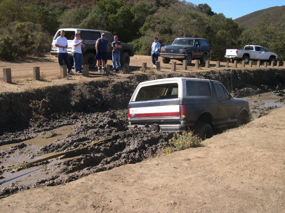 1990 Ford Bronco stuck in mud pit with other vehicles and people nearby