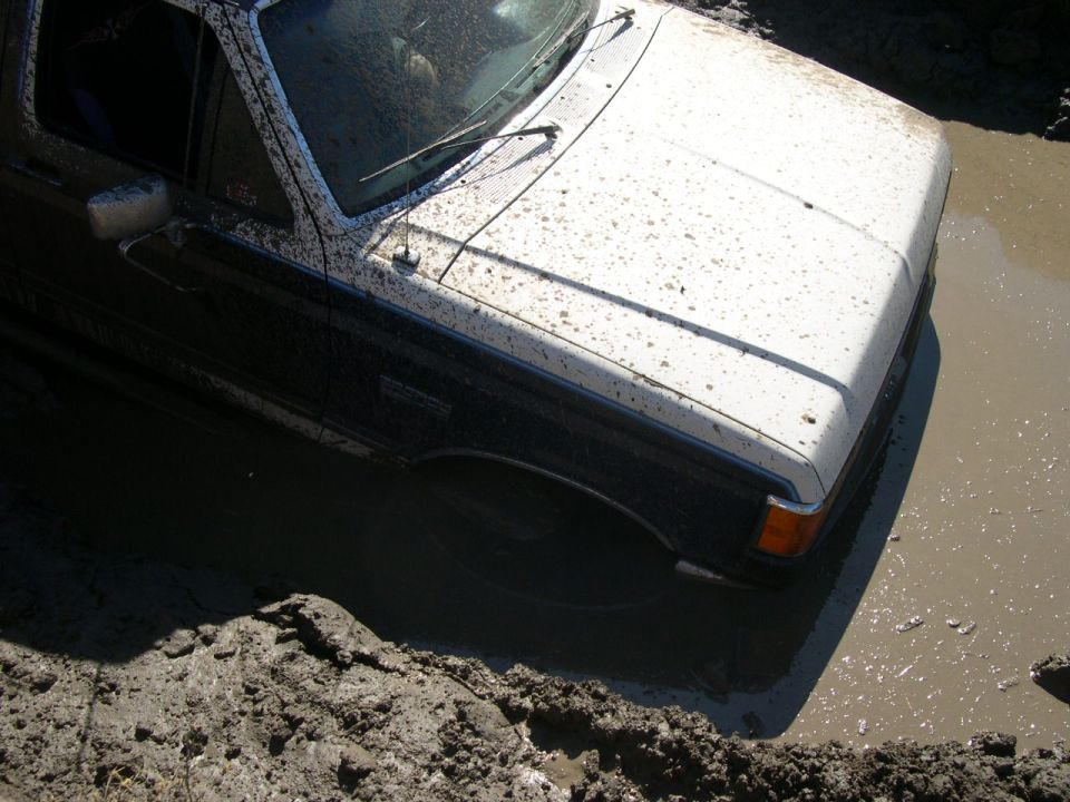 Mud-splattered white and dark blue 1987 Ford Bronco stuck in mud, viewed from the front three-quarter angle