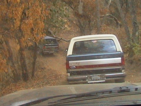 1990 Ford Bronco - Two vehicles drive on a dirt road surrounded by trees with autumn foliage.