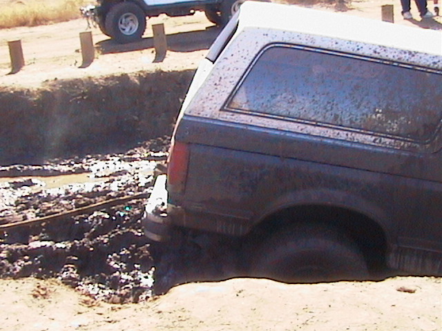 Rear view of a muddy 1990 Ford Bronco stuck in a ditch, with another vehicle visible in the background