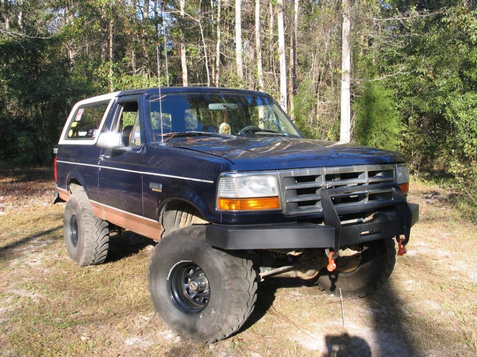 Dark blue Ford Bronco with lifted suspension and large tires parked in a wooded area