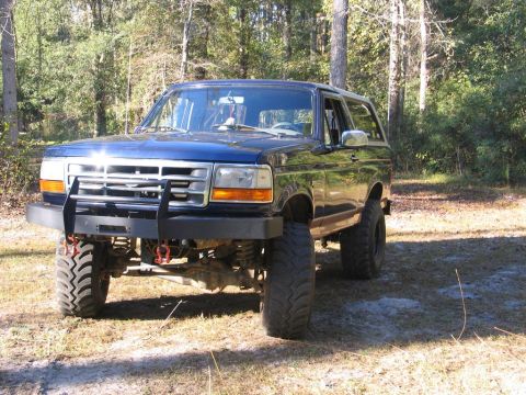 1995 Ford Bronco - This modified Ford Bronco features a custom front bumper and off-road tires, parked in a wooded area.