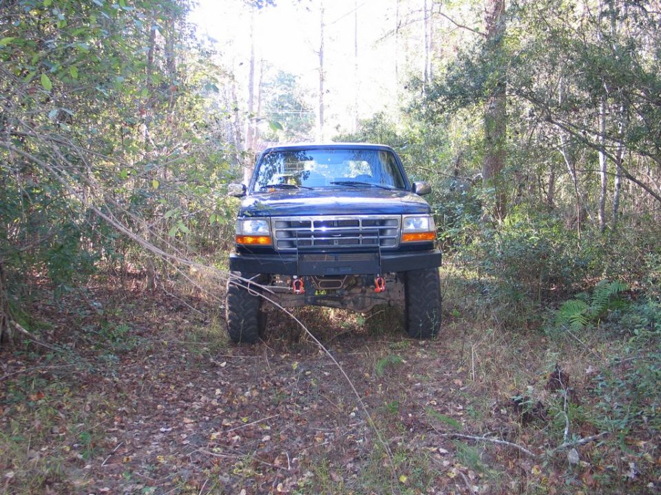 Front view of a dark blue 1995 Ford Bronco with a custom bumper parked on a dirt path in the woods