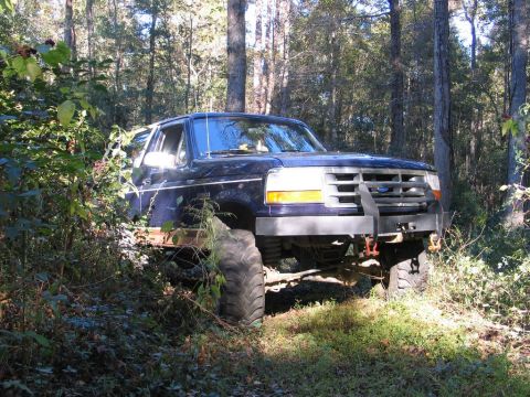 1995 Ford Bronco - A dark blue 1995 Ford Bronco is parked off-road in a wooded area, equipped with large tires and a custom bumper.