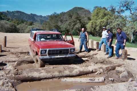 1978 Ford Bronco - A group of men observe a red SUV navigating a challenging muddy obstacle course.
