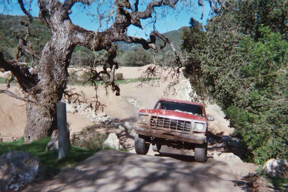 Red 1978 Ford Bronco driving on a dirt path with large trees