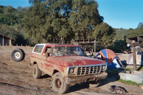 1978 Ford Bronco - Mud-covered 1978 Ford Bronco with a spare tire on a trailer sits at a campsite next to tents.