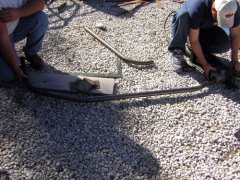 1995 Ford Bronco - Two individuals are working on a bent black car bumper on a gravel lot.