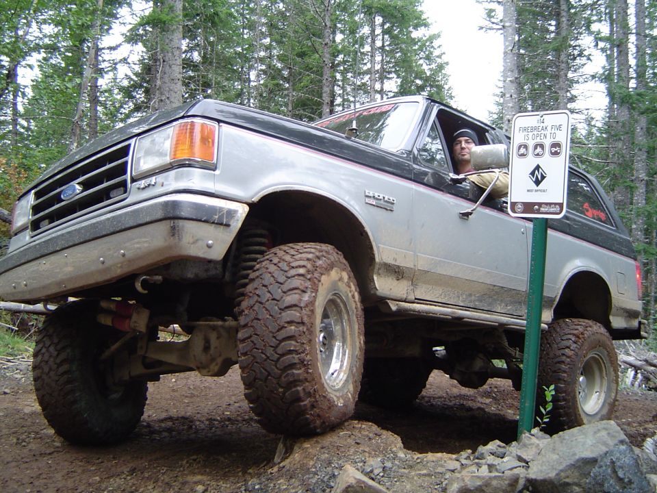 Silver and black lifted Ford 4x4 Bronco with driver flexed over rocks beside a trail sign