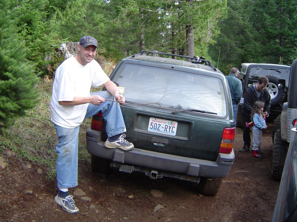 Man in cap and white shirt posing with foot on rear bumper of a green Jeep Cherokee on a trail