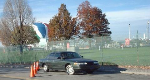1997 Ford Crown Victoria - A 1997 Ford Crown Victoria is parked outside a fenced industrial area with a large spherical building in the background.