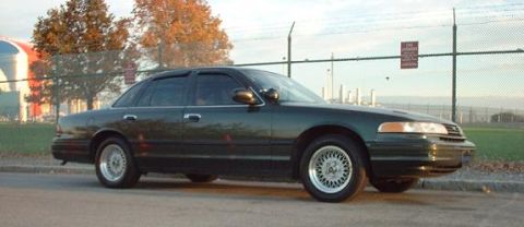 1997 Ford Crown Victoria - A dark green Ford Crown Victoria sedan viewed from the passenger-side front three-quarter, wearing wire-spoke-look wheel covers and parked…