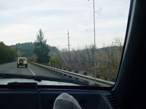 1987 Ford Bronco - A white SUV with a spare tire on the back drives down a highway surrounded by trees and utility poles.