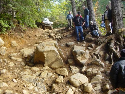1987 Ford Bronco - A 1987 Ford Bronco tackles a challenging, rocky incline on an off-road trail, observed by a group of people.
